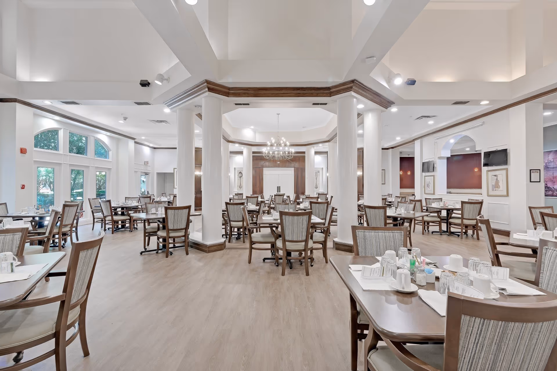 Spacious, well-lit dining room with multiple set tables and chairs, decorative columns, and chandeliers.
