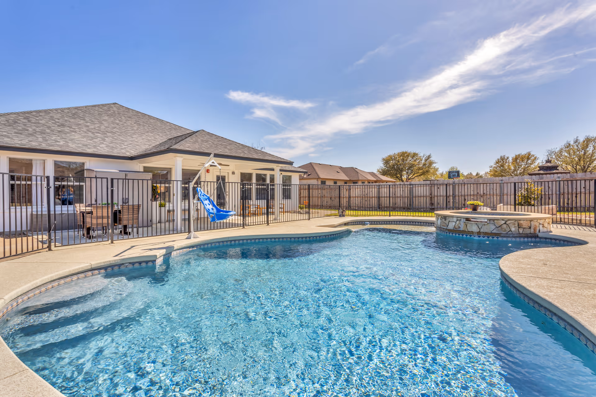 Outdoor swimming pool area with clear blue water, surrounded by a concrete deck and black safety fence. There is a blue pool lift chair for accessibility. In the background, there is a single-story building with a covered patio area furnished with tables and chairs. The sky is clear with some wispy clouds.