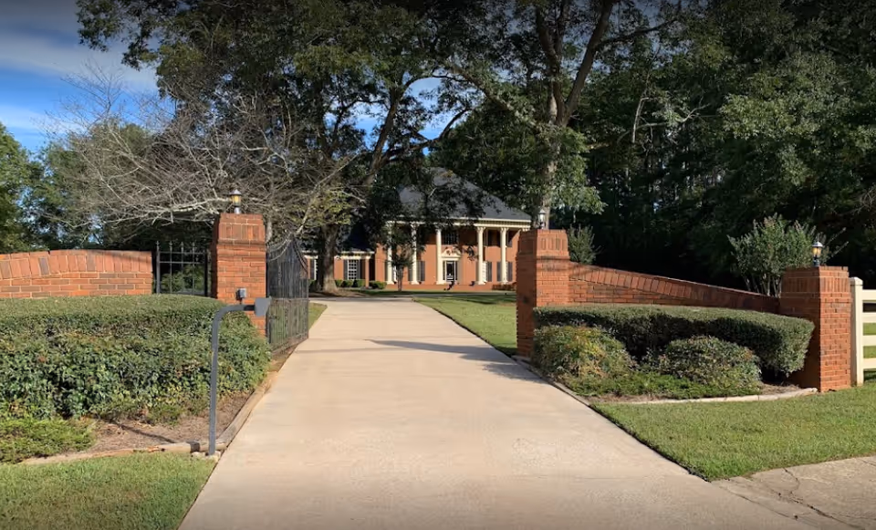 A long driveway leading to a large brick house with white columns, surrounded by green bushes and trees under a clear blue sky.