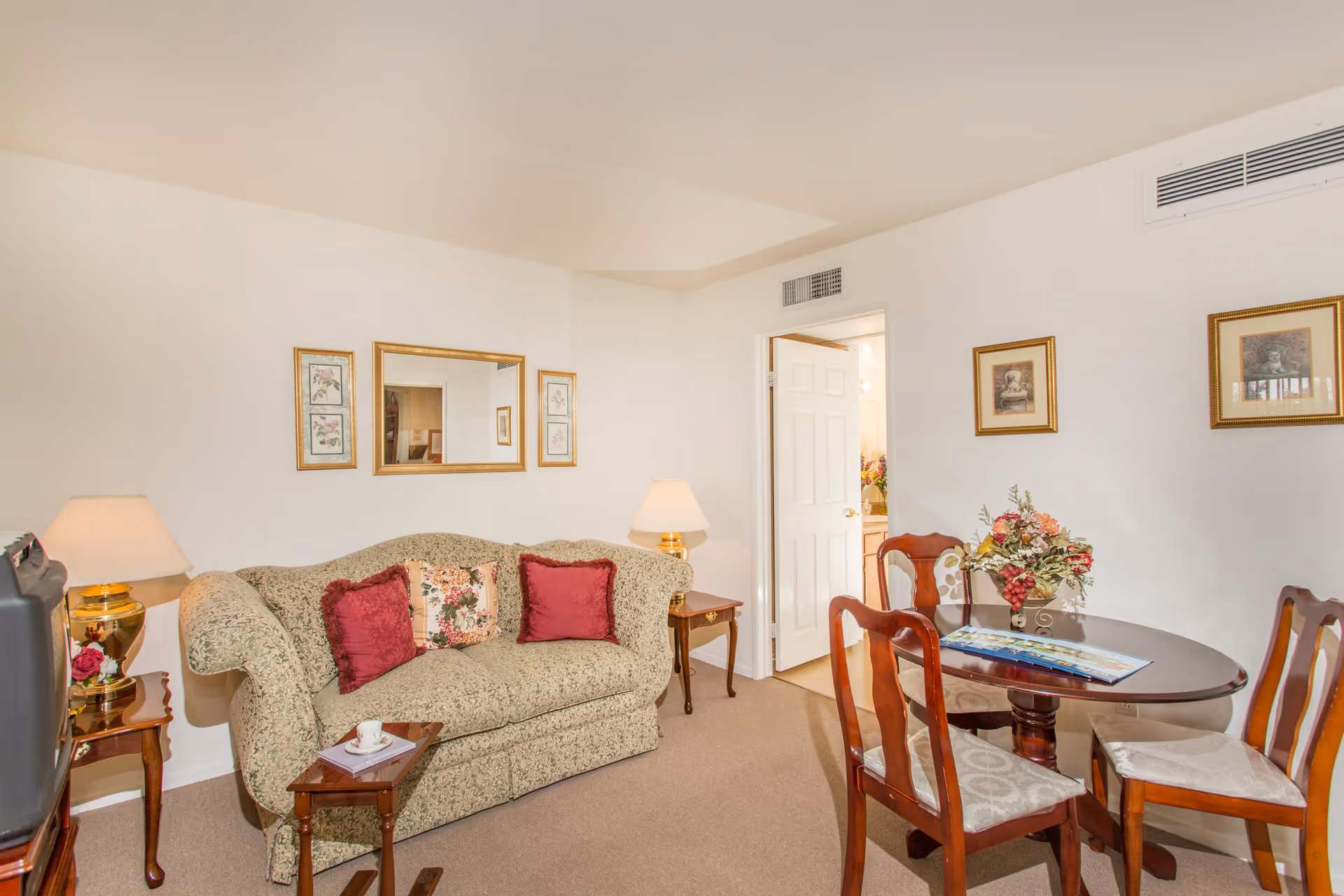 A cozy living room area in a senior facility featuring a patterned sofa with three decorative pillows, two side tables with lamps, a small wooden table with a cup and book, a round wooden dining table with four chairs, floral artwork on the walls, and a doorway leading to another room.