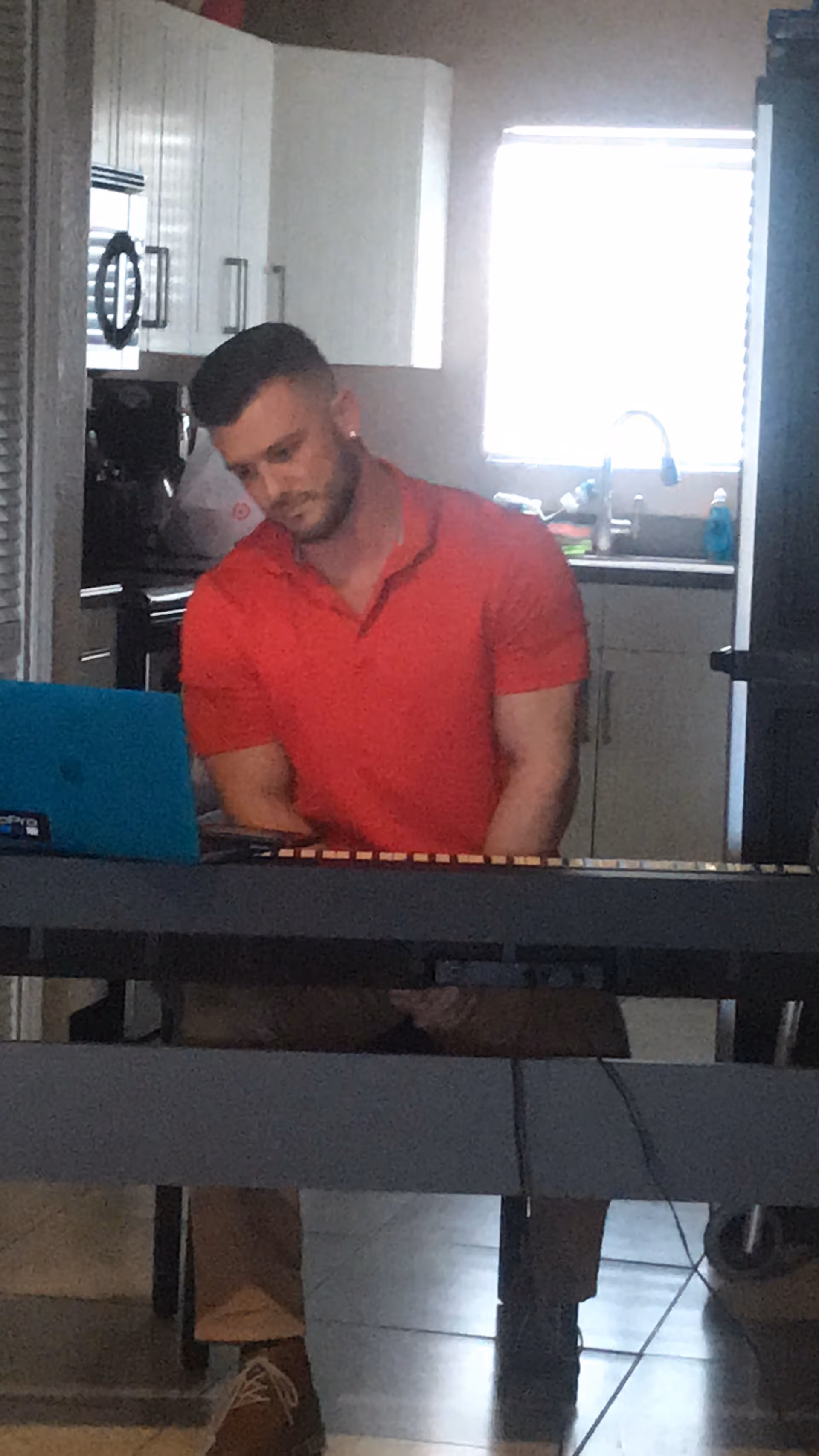A man in a red shirt sitting at a keyboard piano in a kitchen area with white cabinets, a microwave, and a window with blinds.