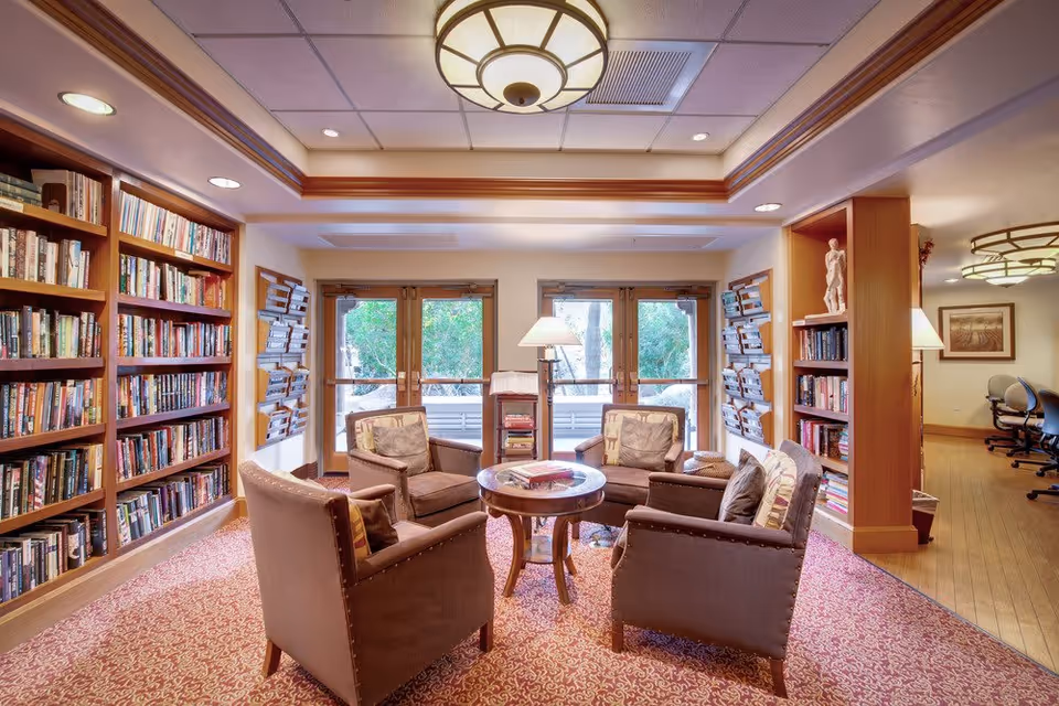 Cozy library room with four cushioned armchairs arranged around a round wooden table. The room features built-in wooden bookshelves filled with books, wall-mounted magazine racks, and large windows letting in natural light. A floor lamp stands near the windows, and the ceiling has recessed lighting and a decorative light fixture. The carpet has a red and beige pattern.
