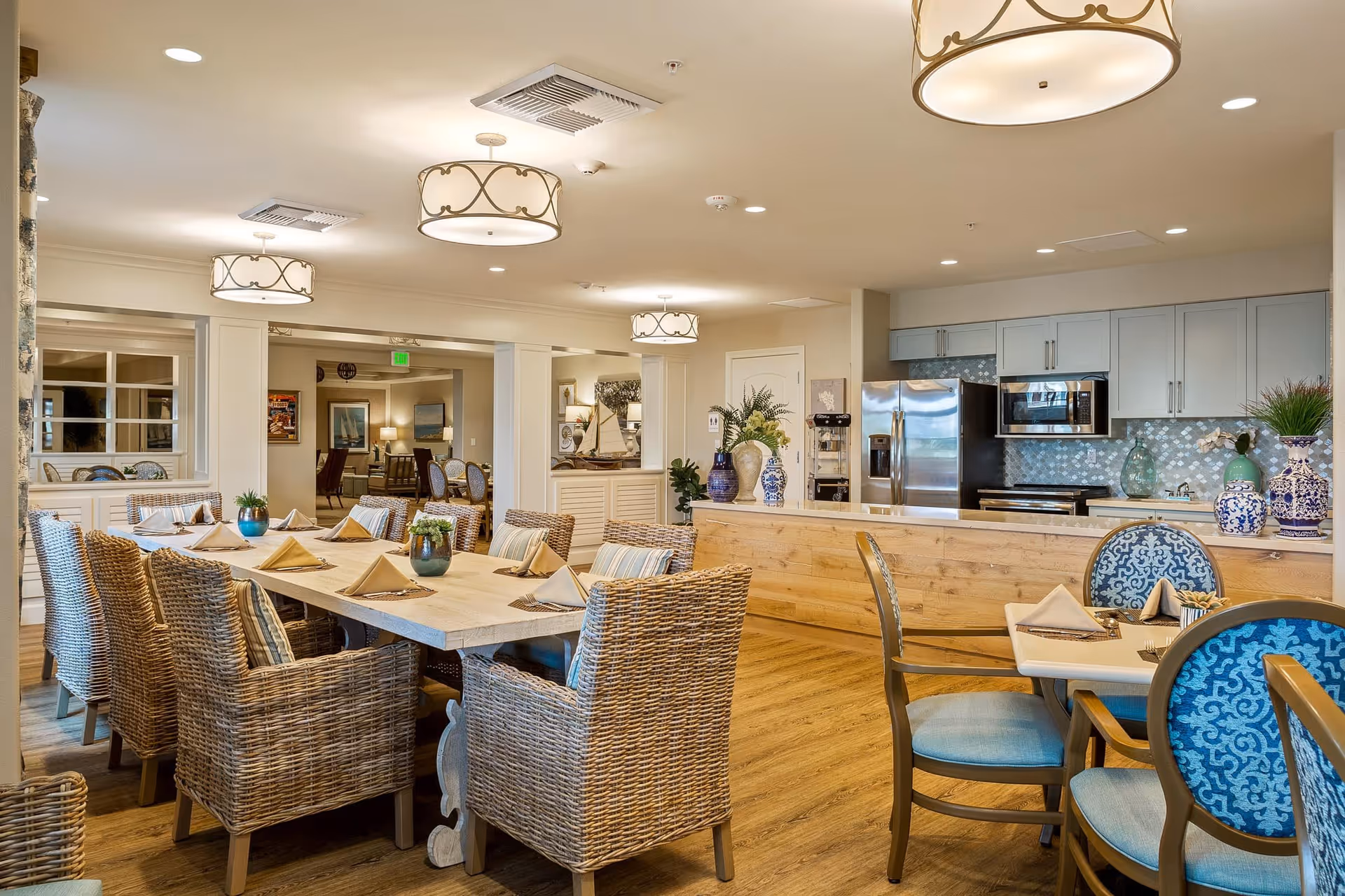 A bright and spacious dining area with a large wooden table surrounded by wicker chairs with cushions. The table is set with folded napkins and small decorative plants. In the background, there is a kitchen area with stainless steel appliances, light blue cabinets, and decorative vases. The room features warm wooden flooring and elegant ceiling lights.