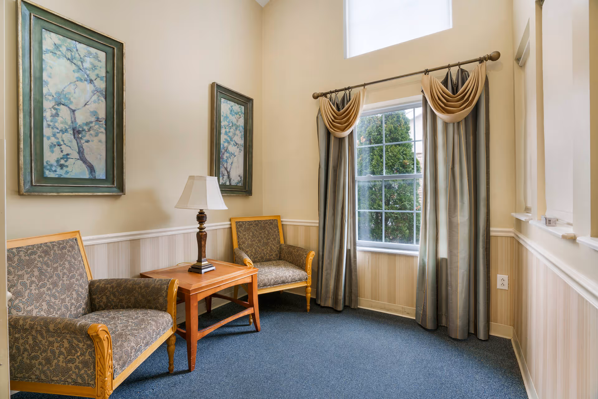 A small sitting area with two patterned armchairs and a wooden side table with a lamp between them. The walls are light beige with white wainscoting, and there are two framed paintings of tree branches on the wall. A window with long curtains and a valance lets in natural light, showing greenery outside.