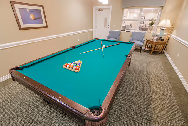 A room with a green felt pool table set up for a game with a triangle rack of billiard balls and two crossed pool cues on the table. The room has beige walls, carpeted floor, two blue armchairs, a wooden side table with a lamp and plant, and a framed picture of a billiard ball on the wall.