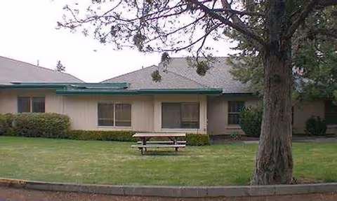 Single-story rehabilitation center exterior with a lawn, tree, and picnic table.