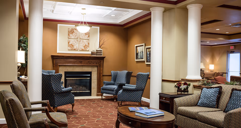 A cozy living room area in a senior living facility with a fireplace centered on a brown wall. The room features four blue patterned armchairs arranged around the fireplace, a patterned sofa with blue cushions, a wooden coffee table with books on it, and two large white columns. The walls are painted in warm tones and decorated with framed artwork. Soft lighting creates a welcoming atmosphere.
