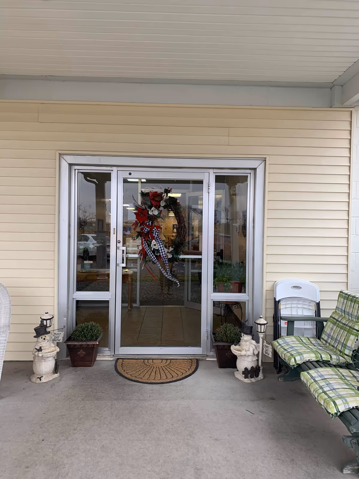 Entrance to a building with glass double doors decorated with a holiday wreath. On either side of the doors are potted plants and snowman decorations holding signs that say 'Just Say Snow'. To the right, there is a green bench with plaid cushions and folded white chairs stacked behind it. The exterior wall is beige with horizontal siding.