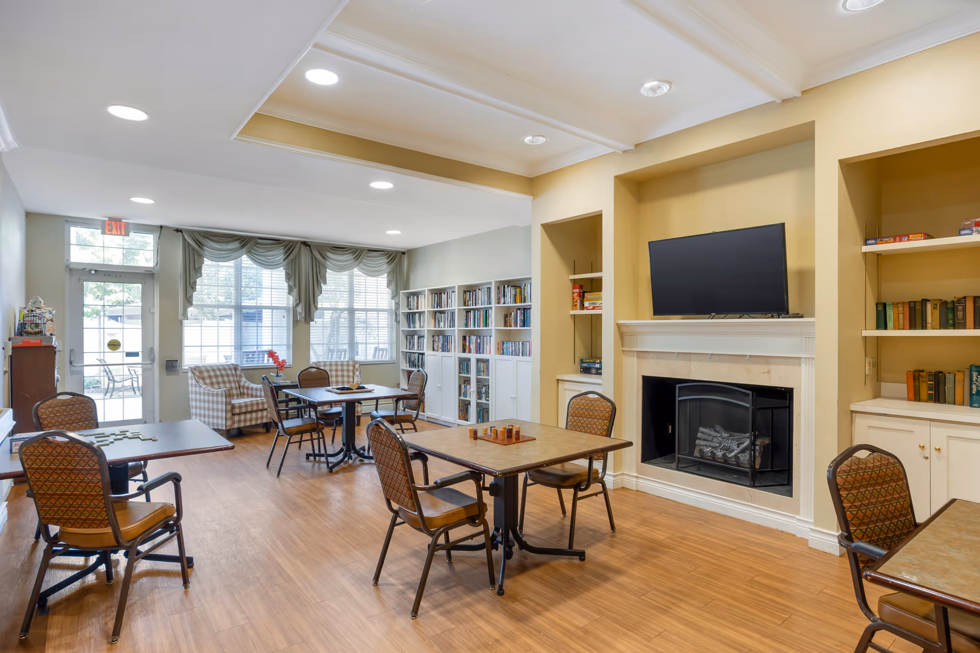 Bright communal living room with tables and chairs, bookshelves, a fireplace and TV near large windows.