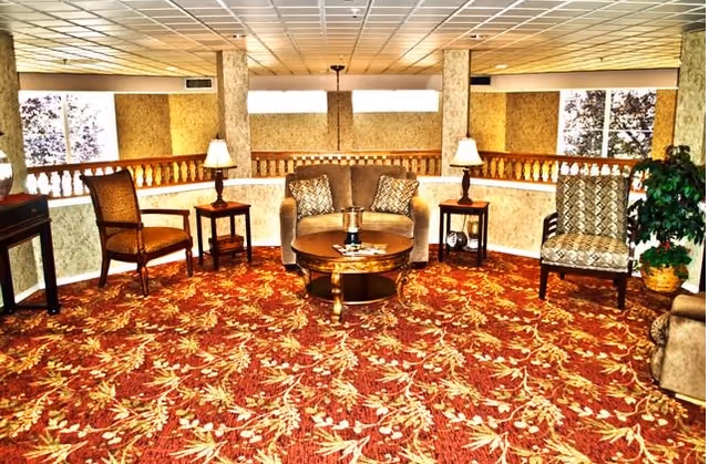 A cozy sitting area with a patterned red carpet, a loveseat with two decorative pillows, two armchairs, two side tables each with a lamp, a round wooden coffee table, and a potted plant near a window.