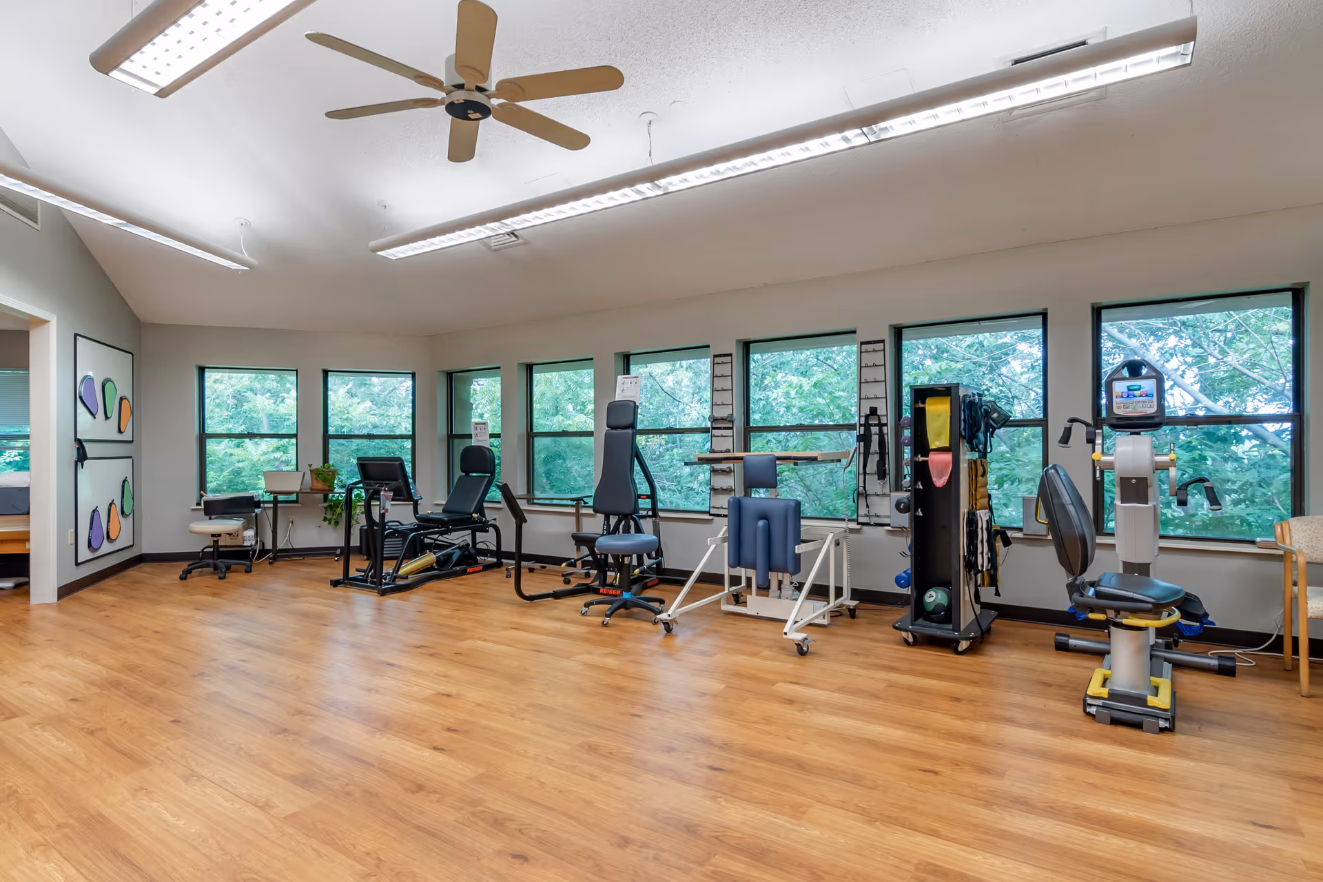 Spacious rehabilitation exercise room with large windows, wood flooring, ceiling fan, and various therapy and exercise machines.
