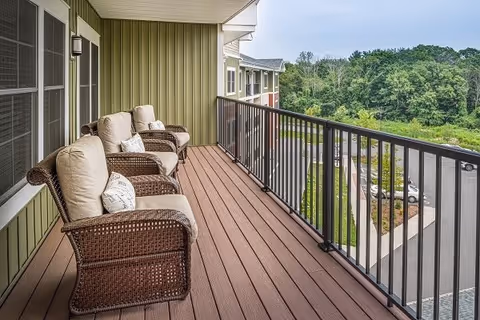 A balcony with three cushioned wicker chairs arranged along the side, overlooking a parking lot and green trees in the distance. The balcony has a brown wooden floor and a black metal railing.