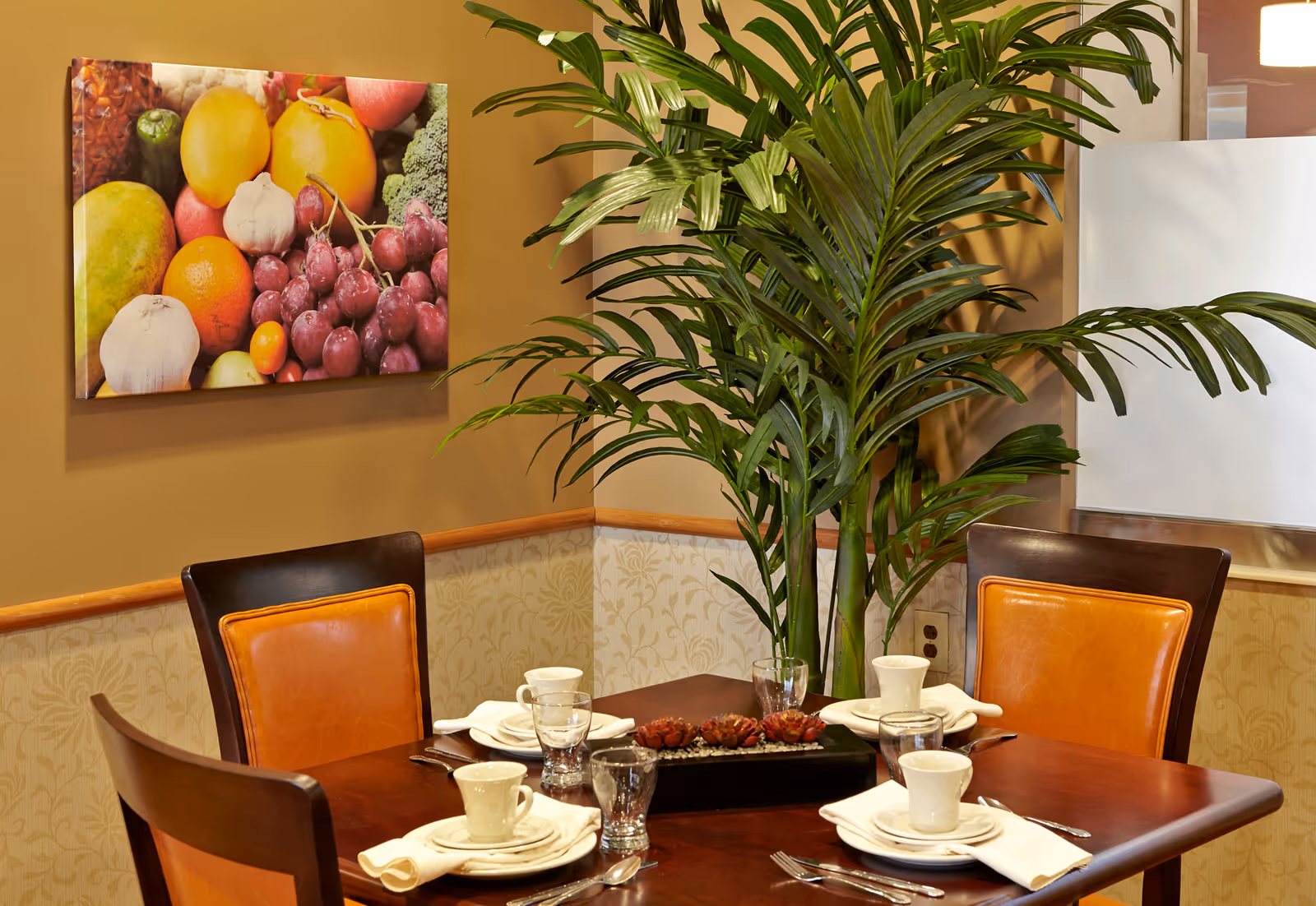 A dining table set for four with white cups, plates, napkins, and silverware. The table has a centerpiece with decorative items and is surrounded by four chairs with brown and orange cushions. A large green plant is placed in the corner, and a colorful fruit-themed painting hangs on the wall behind the table.