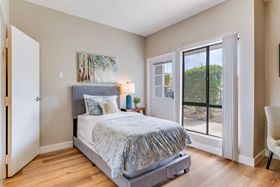 A bright bedroom with a single bed featuring a gray upholstered headboard and patterned bedding. There is a wooden nightstand with a blue lamp and a small plant next to the bed. A colorful painting of trees hangs on the beige wall above the bed. A door is open on the left side, and a large window with vertical blinds and a glass door provide a view of greenery outside. A patterned chair is partially visible on the right side.