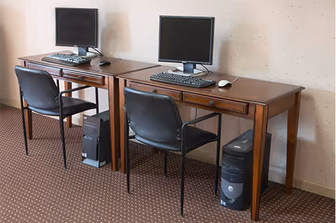 Two wooden desks each with a computer monitor, keyboard, mouse, and a black office chair in front. Two desktop computer towers are placed on the floor under the desks. The room has a carpeted floor and a plain light-colored wall.