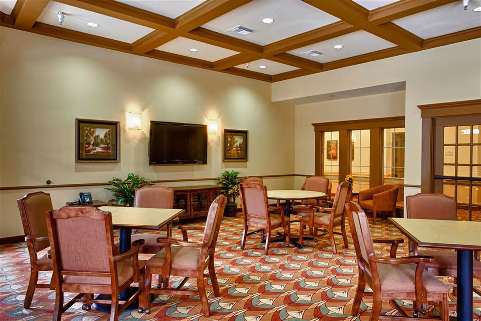 Well-lit communal dining/activity room with multiple tables and upholstered chairs, a wall-mounted TV, framed art, and patterned carpet.
