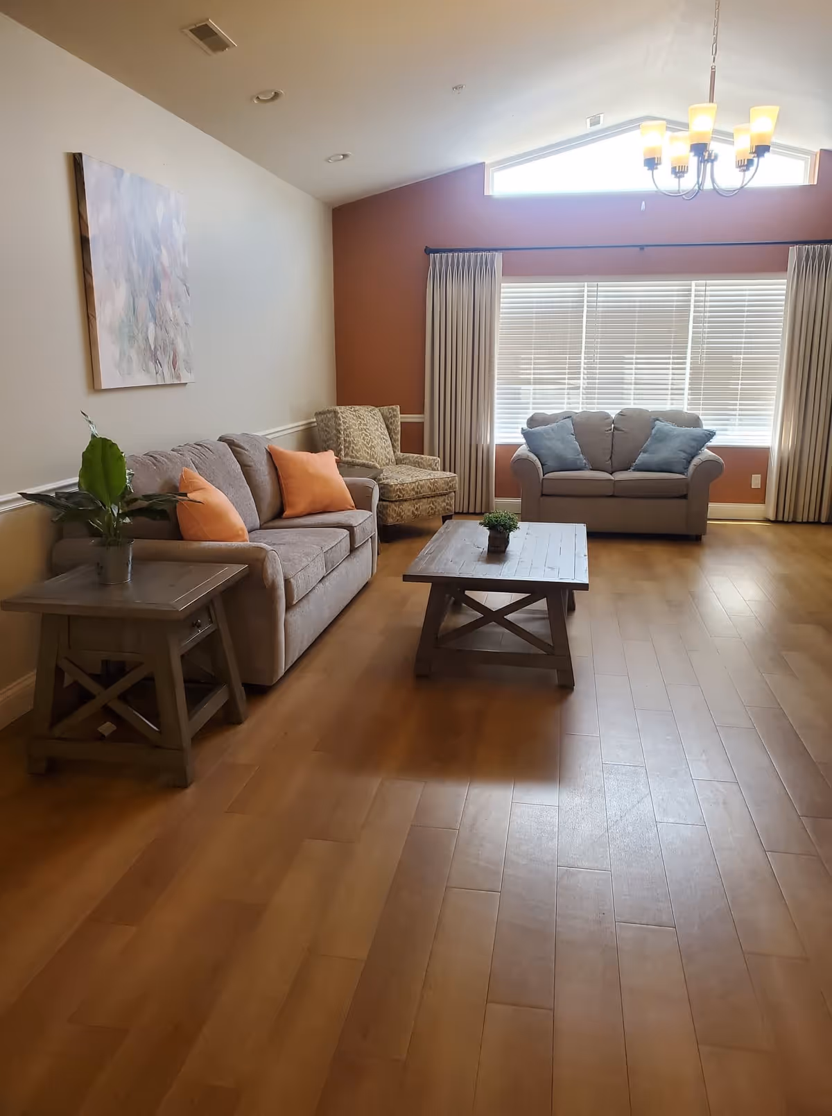 A cozy living room with wooden flooring, featuring a gray sofa with orange pillows, a patterned armchair, a loveseat with blue pillows, a wooden coffee table with a small plant, and a side table with a potted plant. The room has a large window with blinds and curtains, a chandelier hanging from the ceiling, and a piece of abstract artwork on the wall.
