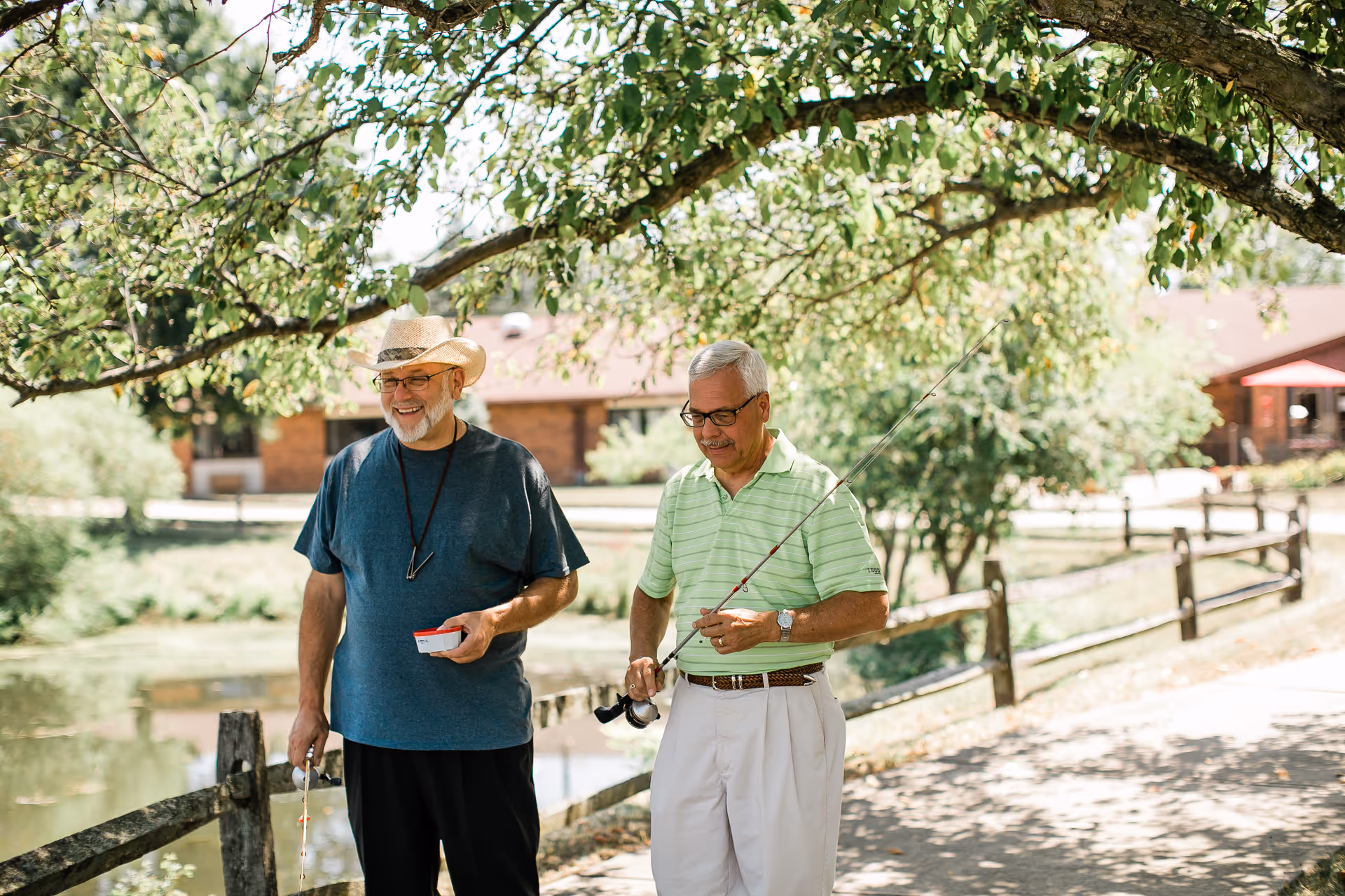 Two elderly men standing outdoors near a wooden fence and a pond, one wearing a straw hat and blue shirt holding a fishing bobber, the other in a light green striped polo shirt holding a fishing rod, with trees and a building in the background.