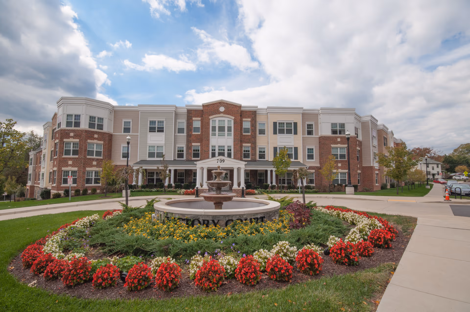 Front exterior view of Victory Court Senior Apartments, a three-story building with a mix of brick and light-colored siding. In front of the building is a circular flower bed with red, white, and yellow flowers surrounding a tiered water fountain. The sky is partly cloudy.