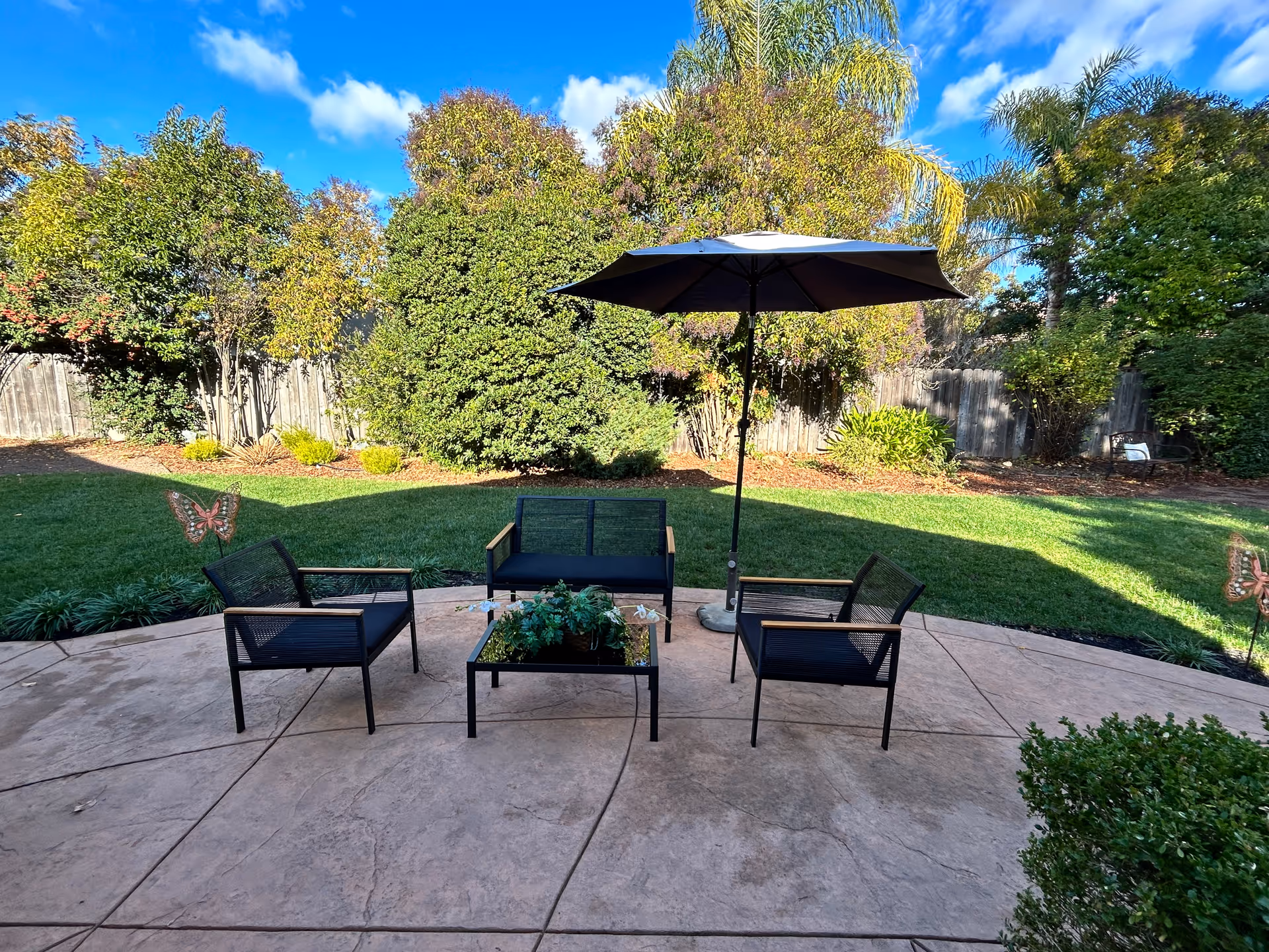 Outdoor patio area with a round concrete floor, featuring a black umbrella, a glass-top coffee table with a plant, a loveseat, and two chairs. The patio overlooks a green lawn bordered by trees and bushes under a blue sky with some clouds.