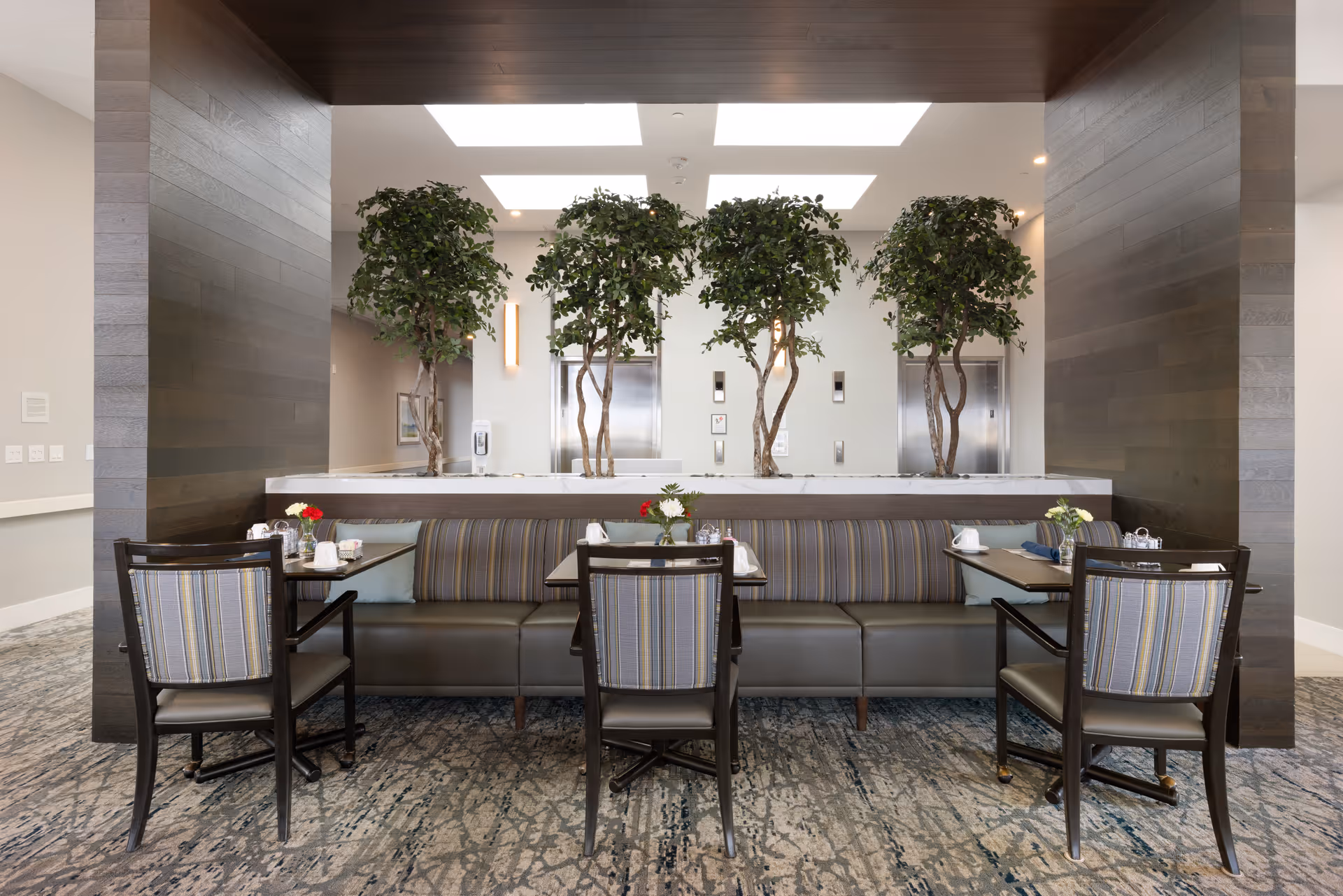 Dining area with banquette seating, small tables and chairs arranged in front of potted indoor trees and elevator doors.