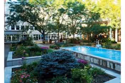 Outdoor courtyard area with a rectangular water feature surrounded by landscaped garden beds filled with various plants and flowers. Trees provide shade over the space, and a multi-story building with large windows is visible in the background.
