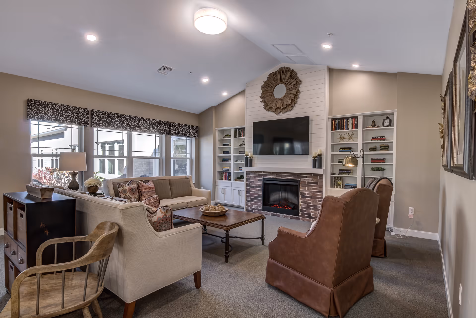 A cozy living room with a beige sofa, two brown armchairs, a wooden coffee table, and a wooden chair. The room features a fireplace with a brick surround and a mounted flat-screen TV above it. Built-in white shelves flank the fireplace, holding books and decorative items. Large windows with patterned valances allow natural light to fill the space. The walls are painted a soft beige, and the ceiling has recessed lighting and a central round light fixture.