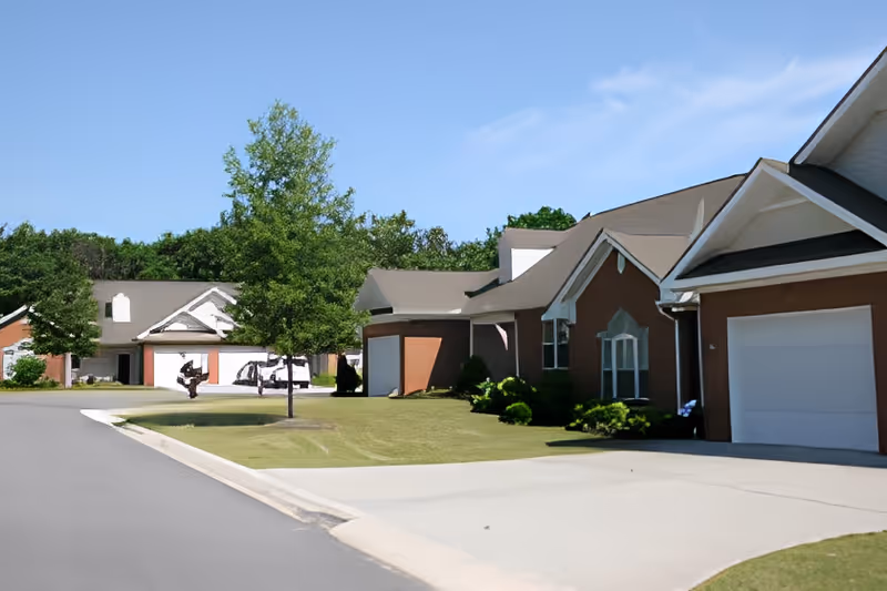 Street view of a residential senior living community with single-story brick cottages, garages, manicured lawns, and trees under a clear blue sky.