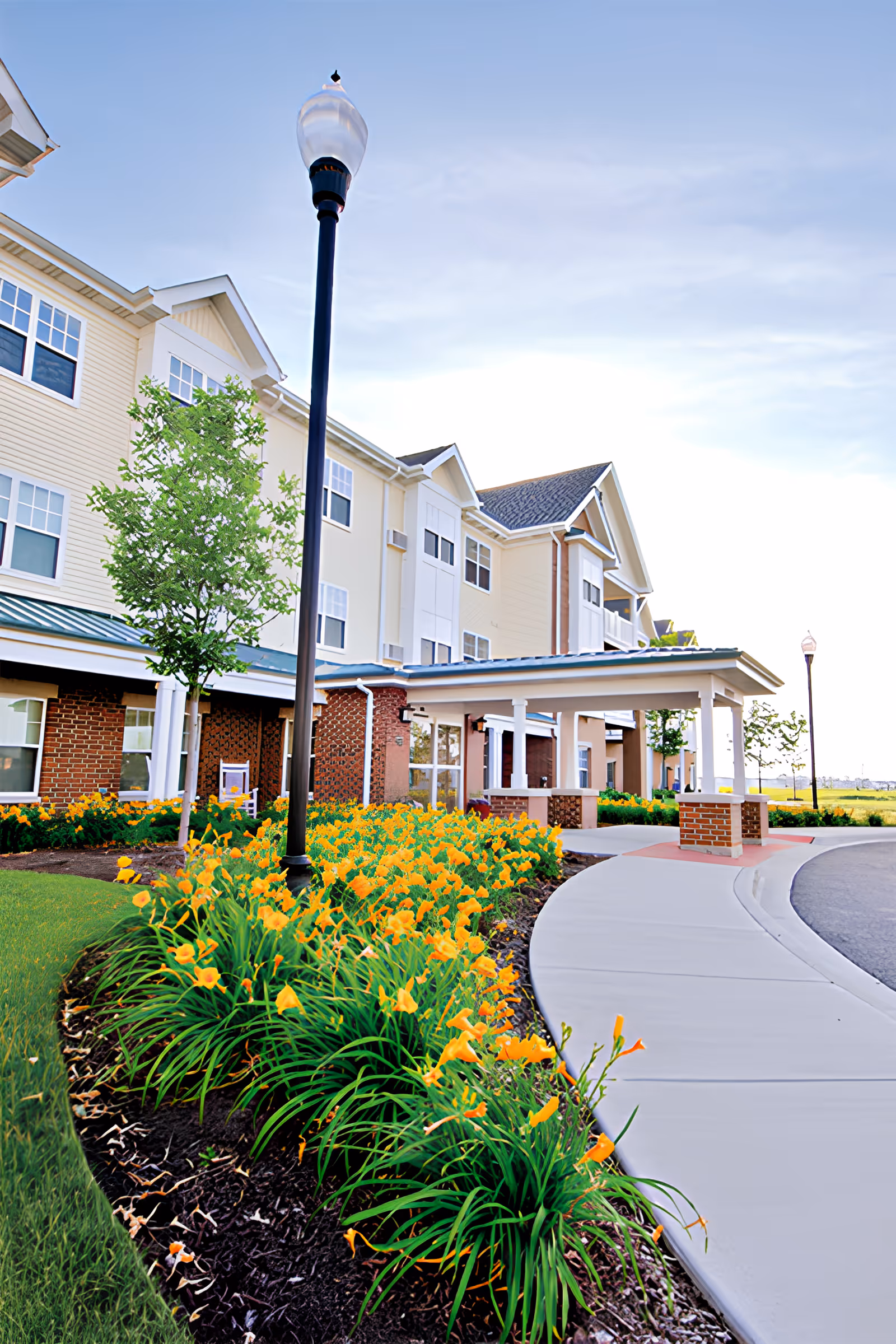 Exterior view of American House Cedarlake showing a three-story building with beige siding and brick accents. There is a covered entrance with white pillars and a curved concrete driveway. Bright orange flowers and green shrubs line the walkway, and a tall black streetlamp stands near the flower bed under a partly cloudy sky.