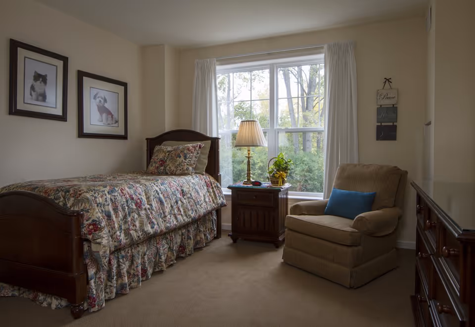 A cozy bedroom with a single wooden bed covered in floral bedding, a wooden nightstand with a lamp and a plant, a comfortable beige armchair with a blue pillow, and a large window with white curtains showing trees outside. Two framed pictures of a cat and a dog hang on the wall.