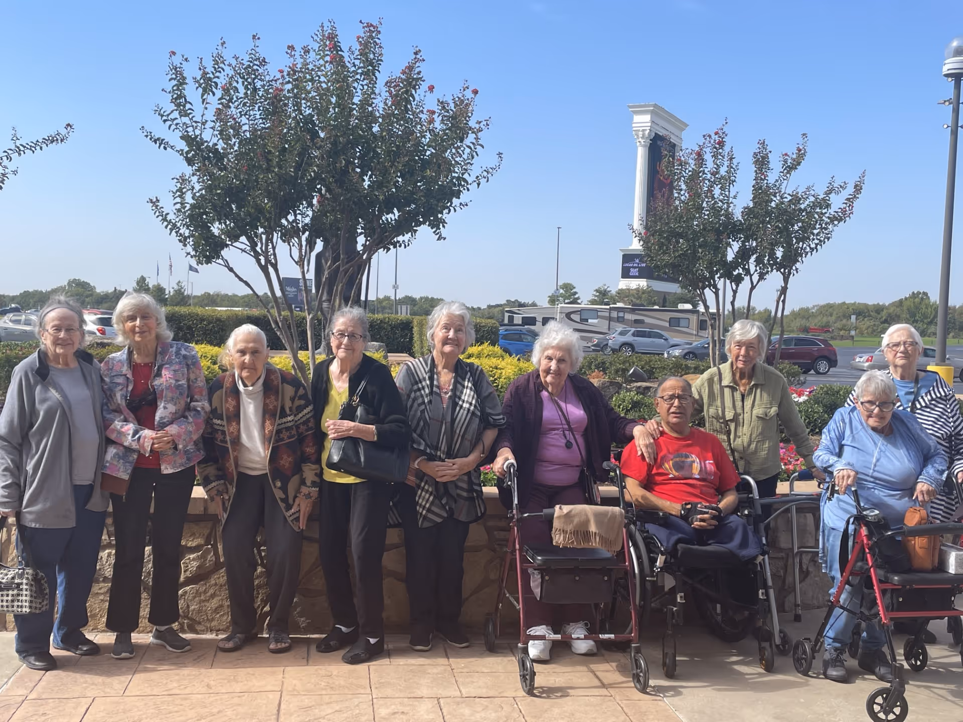 A group of elderly individuals standing and sitting outdoors in front of a low stone wall with trees and a parking lot in the background. Some are using walkers and a wheelchair, and they are smiling and posing for the photo on a sunny day.