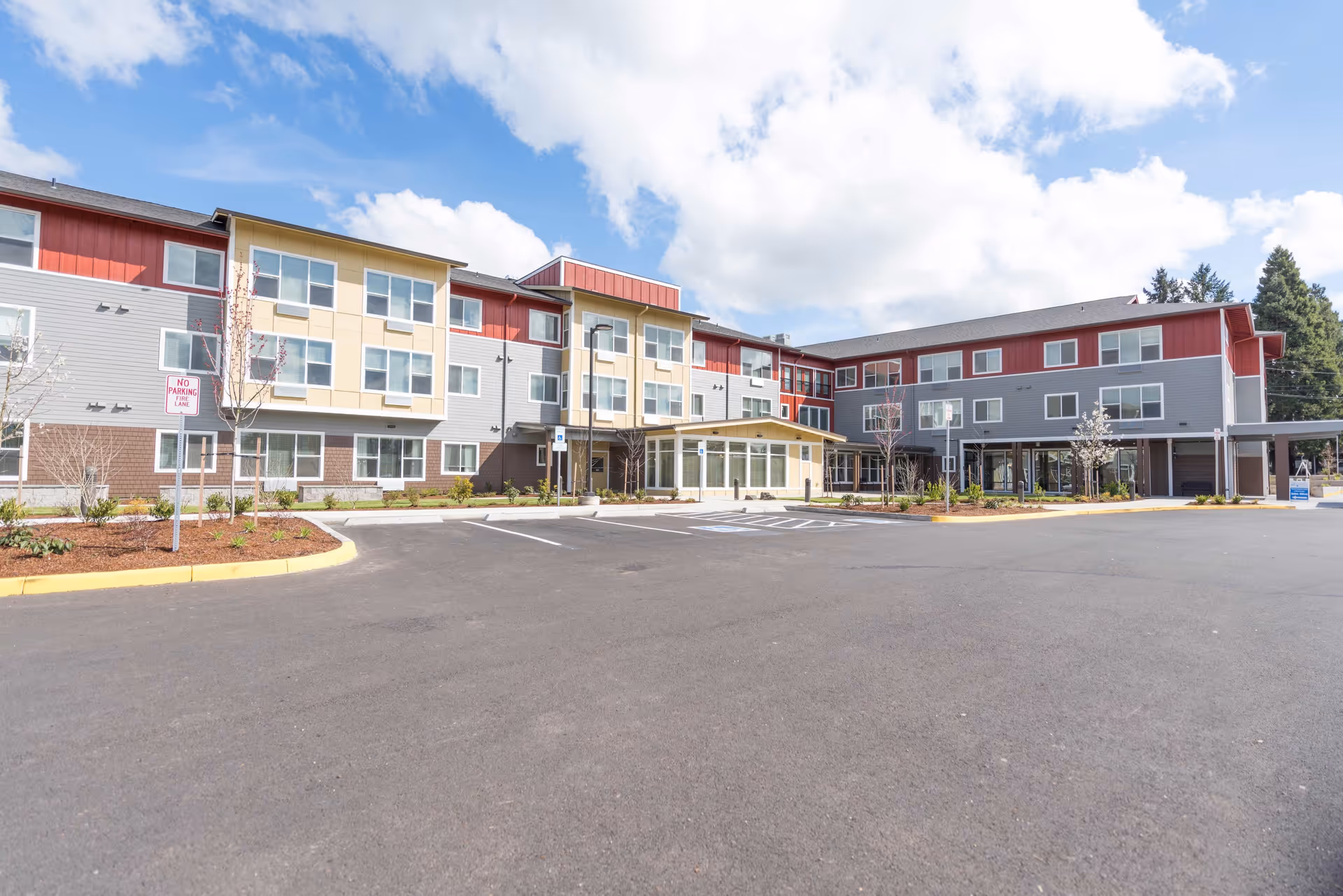 Exterior view of a modern three-story senior living facility named Waverly Place with a large parking lot in front, landscaped areas with small trees, and a partly cloudy sky overhead.