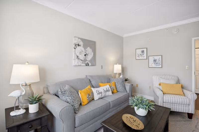 A cozy living room with a gray sofa adorned with decorative pillows, including one that says 'Stay Awhile'. There is a striped armchair with a yellow pillow, two table lamps on either side of the sofa, a dark wooden coffee table with a plant and decorative tray, and floral artwork on the light gray walls.