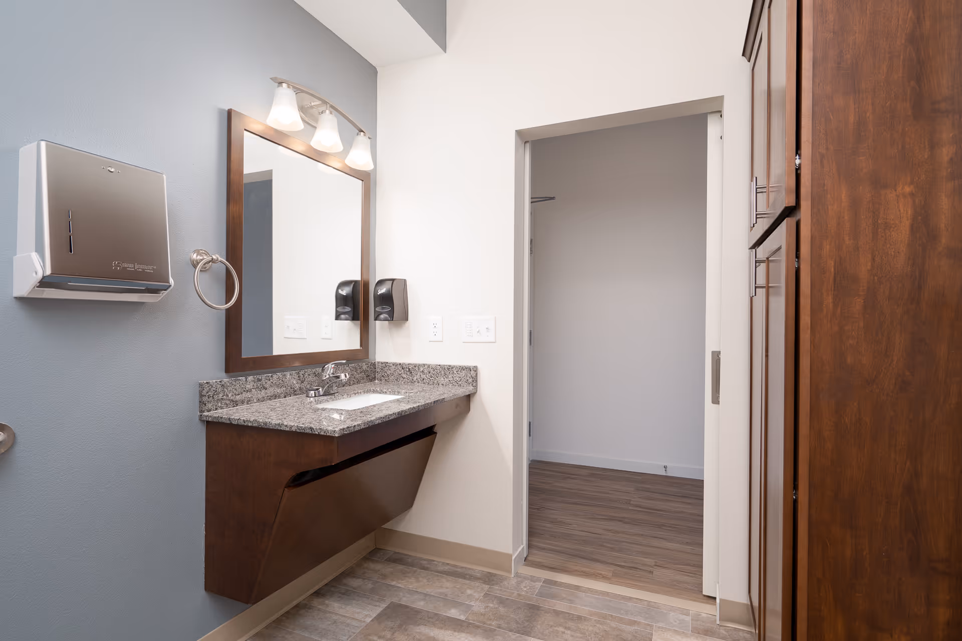 A clean and modern bathroom area with a granite countertop sink, a large mirror with three lights above it, a wall-mounted paper towel dispenser, a soap dispenser, and wooden cabinets. The floor has tile flooring and there is an open doorway leading to a room with wood flooring.