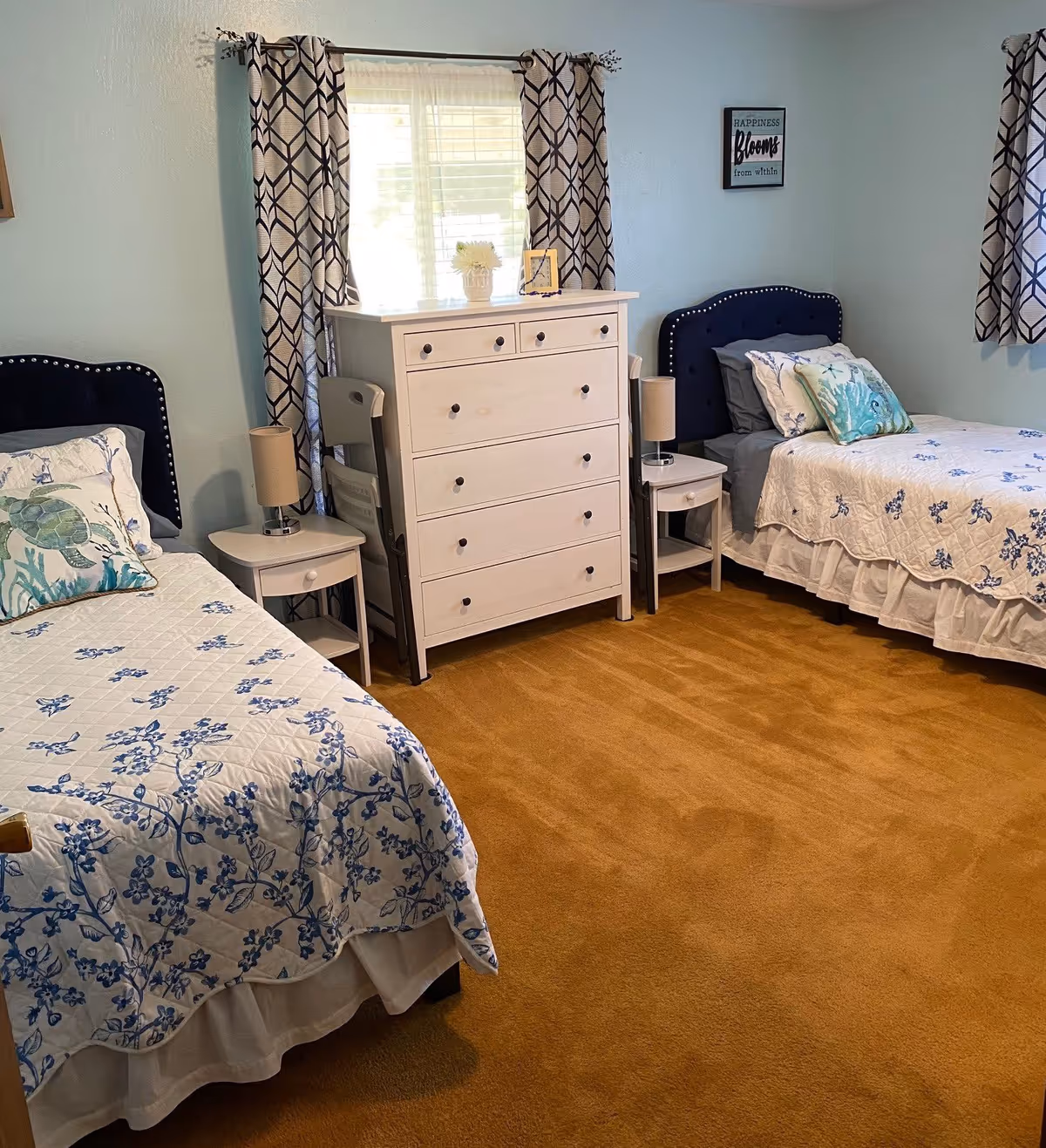 A softly painted bedroom with two twin beds in blue-and-white bedding, a white dresser between them, and patterned curtains at the window.
