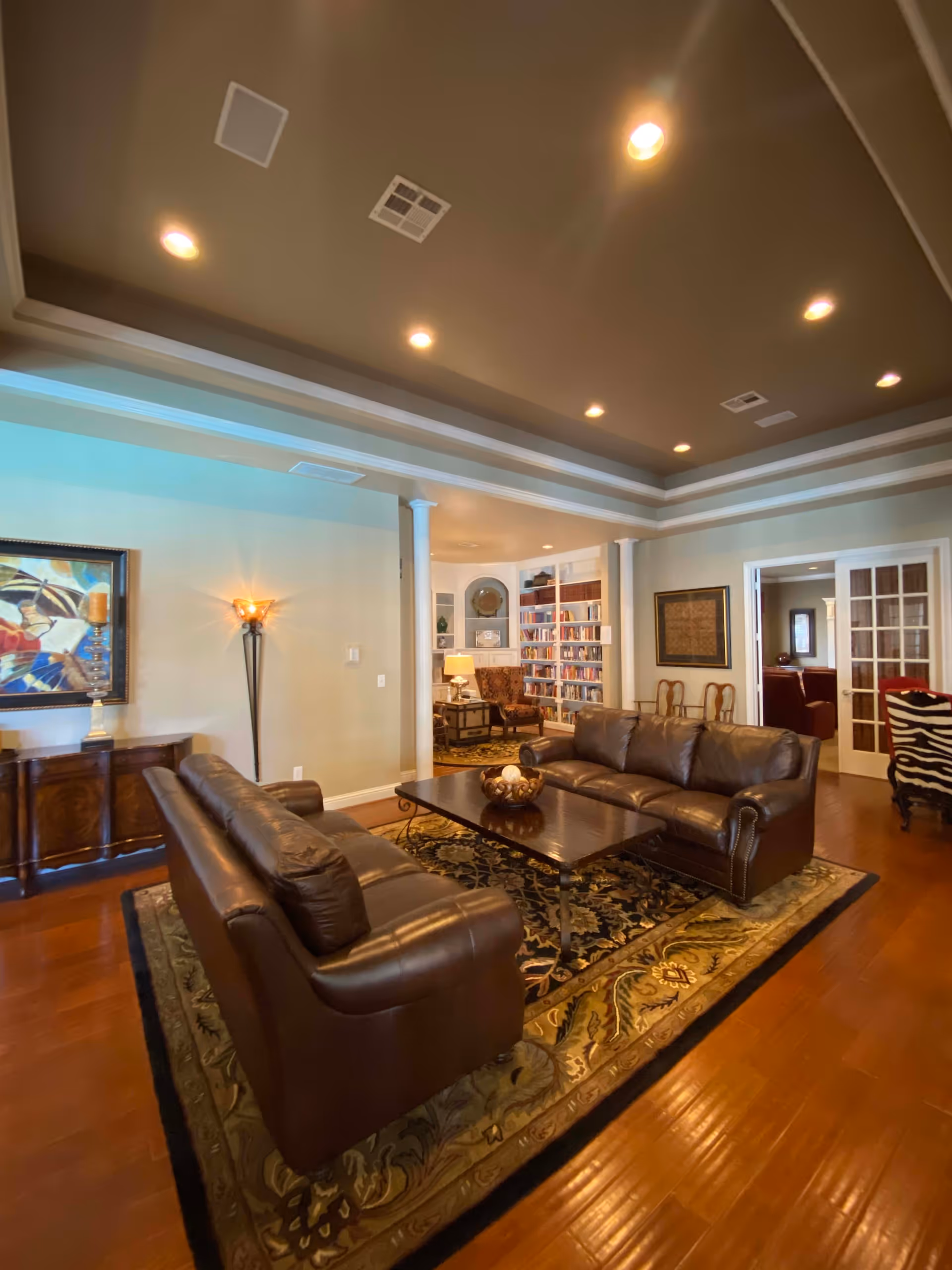 A spacious living room with two brown leather sofas facing each other across a dark wooden coffee table on a patterned area rug. The room features hardwood floors, recessed ceiling lights, and a tray ceiling. In the background, there is a built-in bookshelf filled with books, a side table with a lamp, and additional seating including a patterned armchair. French doors lead to another room with more seating visible.