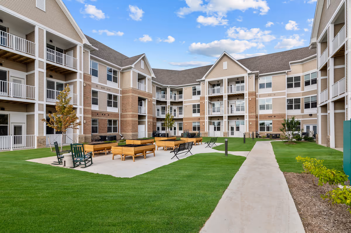 Outdoor courtyard area of a senior living facility with green grass, concrete walkways, wooden planter boxes, benches, and rocking chairs. The courtyard is surrounded by a three-story building with balconies and large windows under a partly cloudy blue sky.