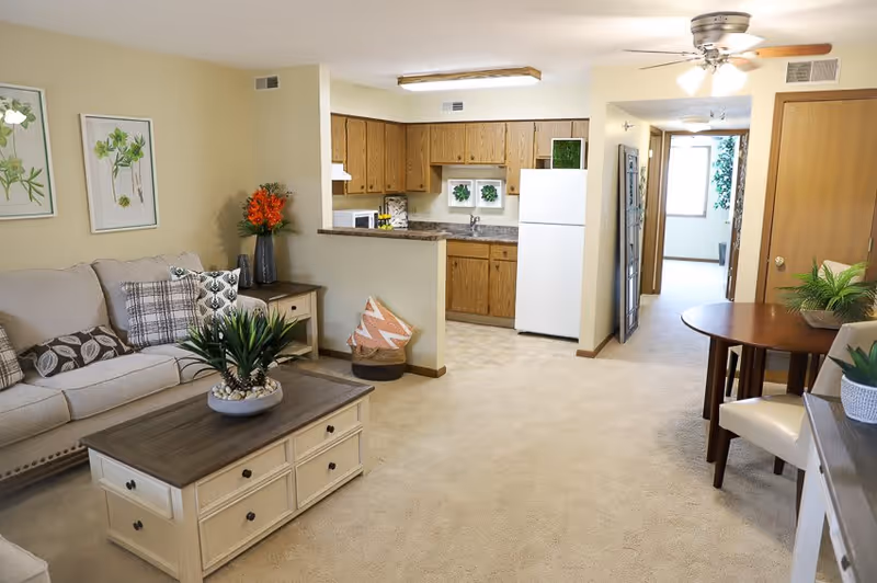 A cozy living area in a senior living facility featuring a beige sofa with patterned cushions, a wooden coffee table with a potted plant, and a small dining table with chairs. The room has beige carpet and walls, with a ceiling fan and light fixture. The kitchen area is visible in the background with wooden cabinets, a white refrigerator, and a countertop. There are framed botanical prints on the wall and decorative plants placed around the room.
