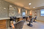 Interior view of a salon area in a senior living facility with three black salon chairs facing three black sinks and mirrors mounted on a tiled wall. The room has wooden cabinetry below the sinks, framed pictures on the walls, and a window letting in natural light.