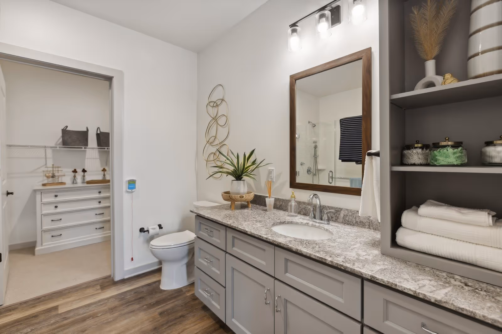 A modern bathroom with a gray granite countertop, an under-mount sink, and a large wooden framed mirror above. To the right, there are open shelves with neatly folded white towels and glass jars containing cotton balls and other toiletries. A toilet is positioned next to the countertop, and a doorway leads to a walk-in closet with white drawers and wire shelving. The floor has wood-like vinyl planks, and the walls are painted white. Decorative elements include a potted plant on the countertop and a metal wall art piece beside the mirror.