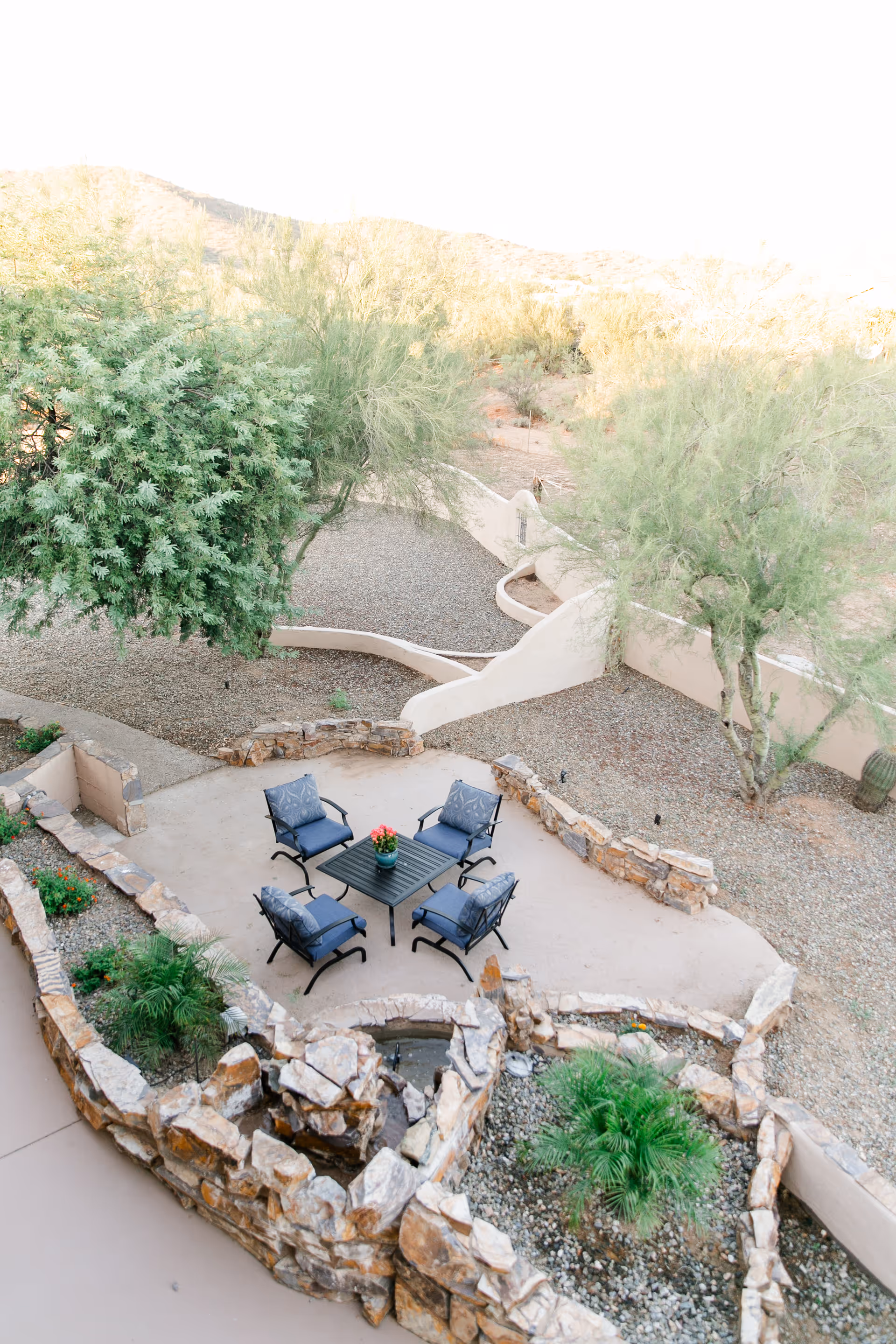 Outdoor patio area with four cushioned chairs arranged around a square table with a small potted plant in the center. The patio is surrounded by low stone walls and landscaping with desert plants and trees. A pathway and additional desert vegetation are visible beyond the patio.
