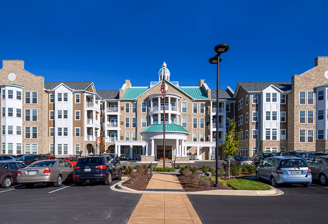 Front exterior view of a large senior living facility building with multiple floors, beige brick and siding, white framed windows, and a central entrance with a green roof and columns. There is a flagpole with an American flag in front, a paved walkway leading to the entrance, and a parking lot with several cars on either side.