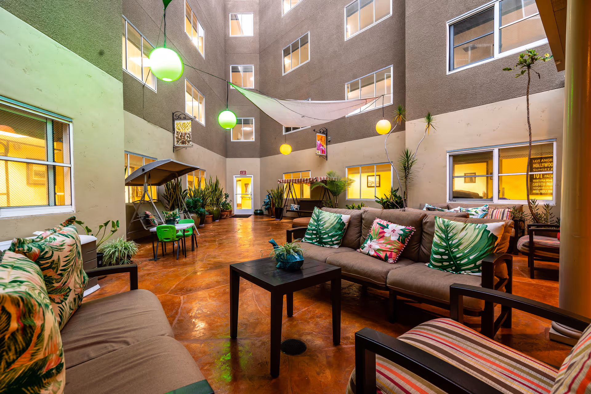 Indoor courtyard area with comfortable seating including sofas and chairs adorned with tropical-themed cushions. The space features hanging spherical lights, potted plants, and a canopy overhead. Windows from surrounding rooms overlook the courtyard.
