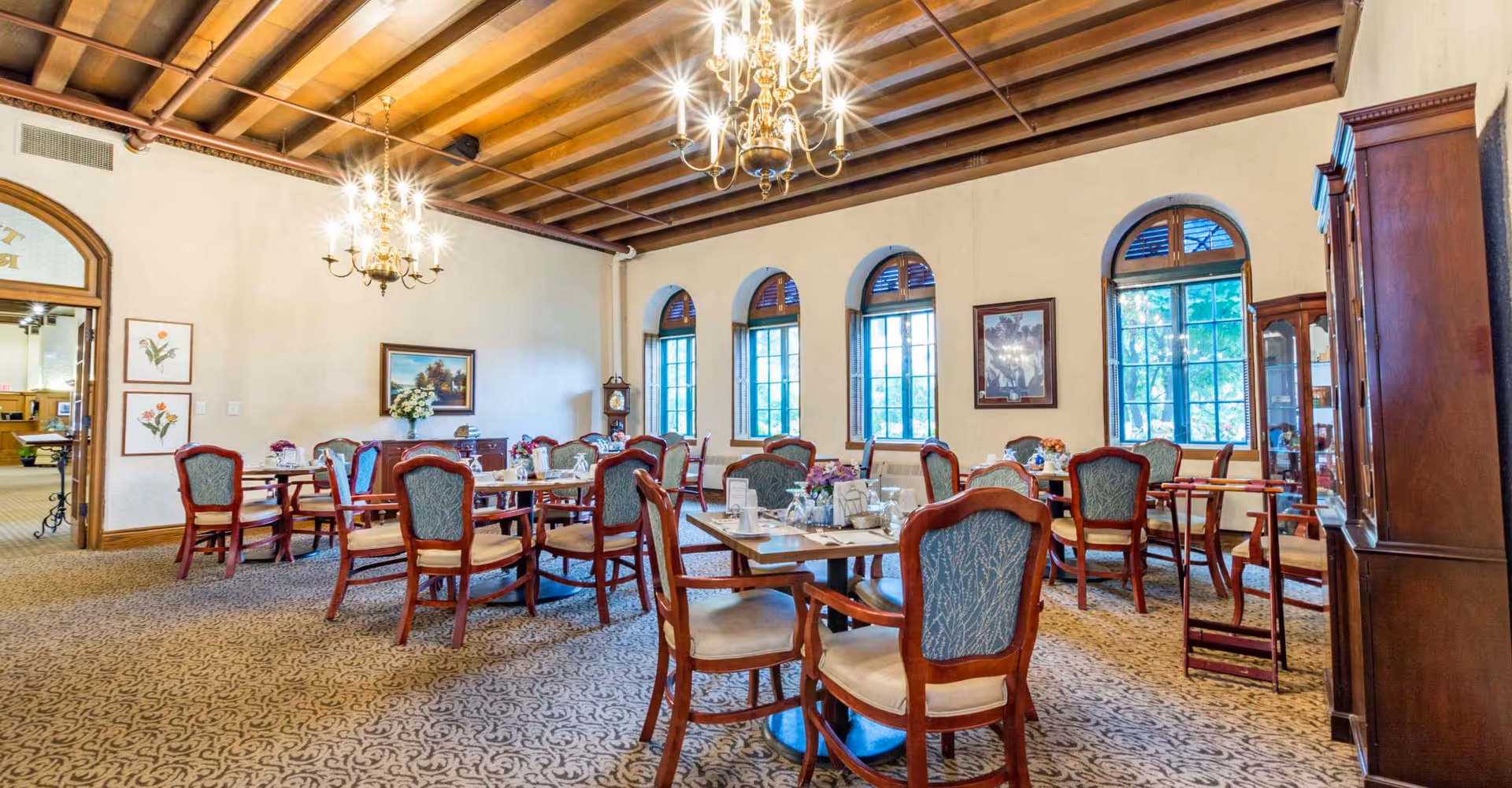 A dining room with multiple wooden tables and chairs featuring blue upholstered backs and beige seats. The room has a patterned carpet, cream-colored walls, and large arched windows letting in natural light. Two ornate chandeliers hang from a wooden beam ceiling. There are framed pictures on the walls and a wooden cabinet on the right side.