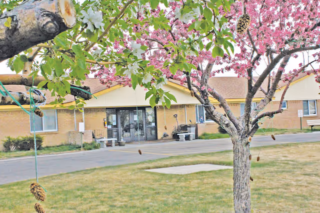 Front entrance of a single-story care facility seen through blossoming trees and a grassy lawn.