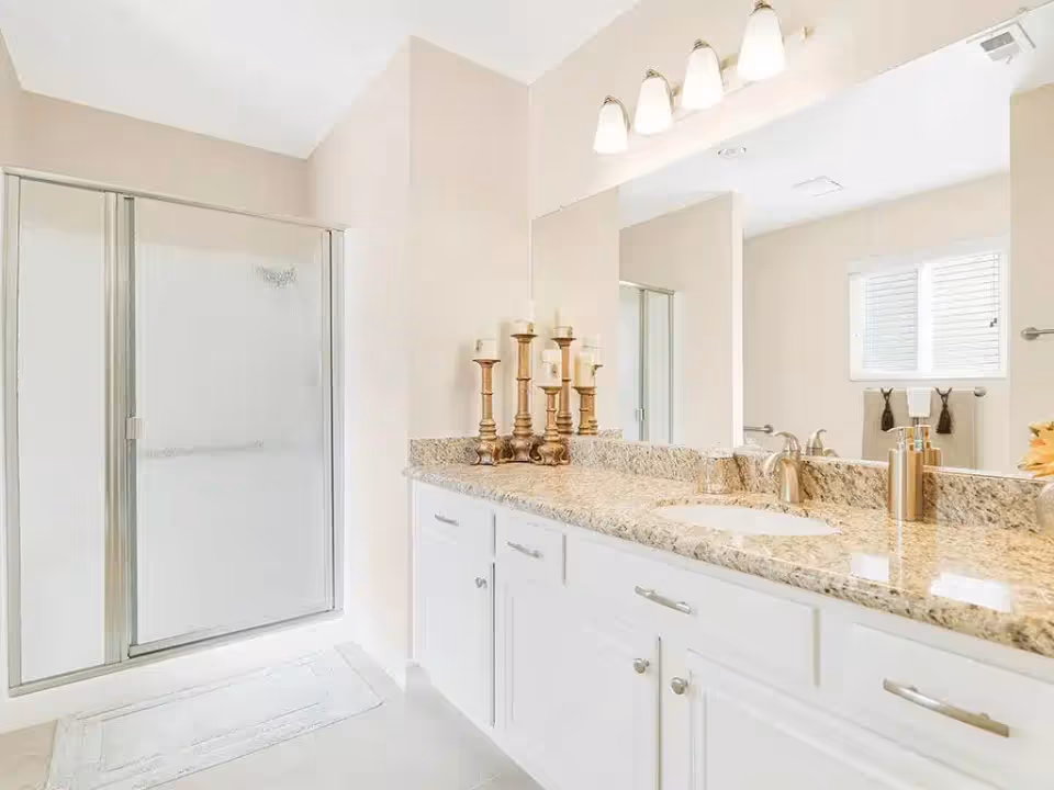Bright bathroom with a granite double-sink vanity, large mirror, glass-enclosed shower, and decorative candle holders.