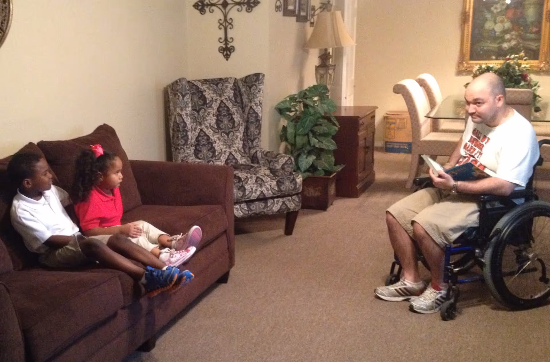 Two children sit on a couch while a man in a wheelchair reads to them in a cozy living room.