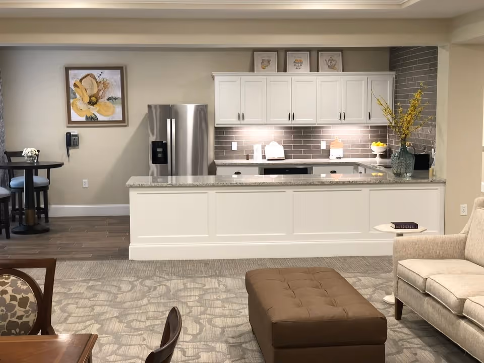 Interior view of a senior living facility kitchen and living area. The kitchen features white cabinets, a stainless steel refrigerator, a gray tiled backsplash, and a granite countertop with a vase of yellow flowers. Adjacent to the kitchen is a small dining area with a round table and two chairs. The living area includes a beige sofa, a brown tufted ottoman, and a small side table with a book on it. The floor has a patterned carpet and wood flooring near the dining area. A framed floral painting hangs on the wall near the dining table.