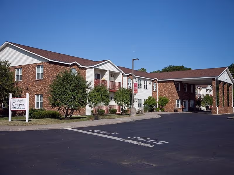Exterior view of The Hearth at Greenpoint senior living facility showing a two-story brick and siding building with a covered entrance, trees, and a parking lot with a stop sign painted on the asphalt.
