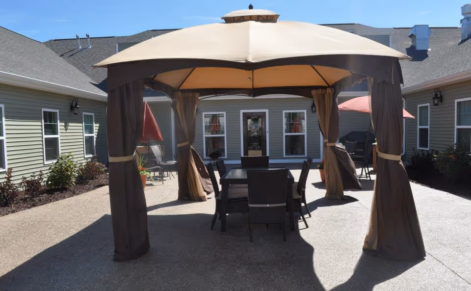 Outdoor patio area at Vicinia Gardens Luxury Retirement Living featuring a large beige and brown canopy with curtains, a black dining table with six chairs underneath, surrounded by a paved courtyard and green building walls with windows.